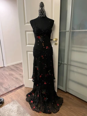 Svart långklänning med blommor och pärlor - Supersnygg svart långklänning. Klänningen har figurnära passform, volangig nederdel och dekorativa pärlor framtill. Den är gudomlig.