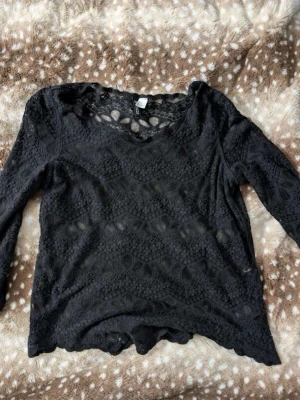 Svart spetsblus med lång ärm - Snygg svart långärmad topp i genombruten spets med vågig kant nertill och vid ärmslut. Rund halsringning och transparent material ger en trendig look. Perfekt att styla lager på lager för en cool outfit.