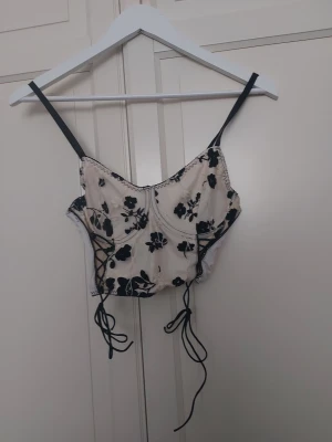 Vit korsett med svarta blommor - Säljer en snygg vit korsett med svarta broderade blommor och svarta banddetaljer. Axelbanden är svarta och korsetten har snörning i sidorna för justerbar passform. Materialet är tunt och genomskinligt med broderade detaljer för en trendig look.