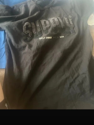 Svart SUPPLY t-shirt med text - Snygg svart t-shirt från SUPPLY med stor text i 3D-effekt på bröstet och mindre tryck under. Klassisk passform och rund halsringning, perfekt till jeans eller shorts. Materialet känns mjukt och bekvämt mot huden.