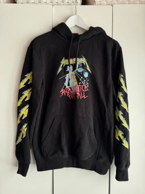 Svart Metallica hoodie. XS. H&M.  - Svart hoodie med Metallica, And Justice For All-tryck på bröstet och blixtmotiv i gult längs båda ärmarna och på ryggen. Storlek XS, (rymlig). Regular Fit, H&M. Mjuk borstad insida. Stor känguruficka framtill och justerbar huva med snören. Perfekt för dig som gillar bandmerch och vill sticka ut med en ikonisk design. Lite använd. Fint skick.