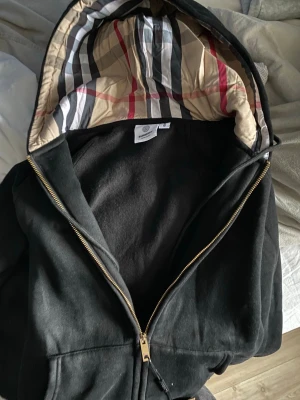 Svart hoodie från Burberry med dragkedja - Säljer en svart hoodie från Burberry med guldfärgad dragkedja och klassiskt rutigt foder i huvan. Hoodien har en avslappnad passform och är tillverkad i mjukt material. Perfekt för dig som vill ha en stilren och lyxig look med ikoniska Burberry-detaljer. Tycker själv den är lite för liten i passformen därför jag ej kan ha den även om jag är M vanligtvis, finns även i grå