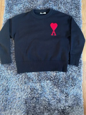 Svart stickad tröja från Ami Paris - Svart stickad tröja från Ami Paris med röd hjärtlogga och bokstaven A broderad på bröstet. Tröjan har rund halsringning, ribbade muddar och nederkant. Perfekt för dig som gillar stilrena plagg med en twist.