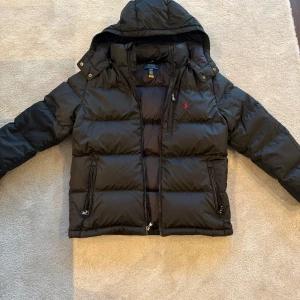 Svart pufferjacka från Polo Ralph Lauren - Svart pufferjacka från Polo Ralph Lauren med huva och dragkedja framtill. Jackan har två fickor med dragkedja och den klassiska röda Polo-loggan på bröstet. Perfekt för kalla dagar och riktigt snygg till streetwear-stilen.