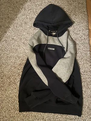 Grå och marinblå hoodie från Nicce - Snygg hoodie från Nicce i marinblått och grått med kontrastpaneler och broderad logga på bröstet. Klassisk huva med dragsko och ribbade muddar vid ärmslut och nederkant. Perfekt för en chill och sportig look. Lite små skador på armen .