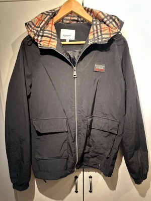 Svart Burberry vindjacka med huva - Stilren svart vindjacka från Burberry med klassiskt rutigt foder i huvan. Jackan har dragkedja framtill, två stora fickor med lock och justerbar dragsko i nederkant. Märkespatch på bröstet och elastiska muddar vid ärmsluten. Perfekt för en trendig streetwear-look.