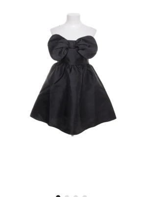 klänning med rosett Only  - Supersnygg svart axelbandslös kortklänning med stor rosett framtill. Klänningen har en utställd form och är gjord i ett glansigt material som ger en lyxig känsla. Perfekt för dig som vill sticka ut och känna dig extra fin.