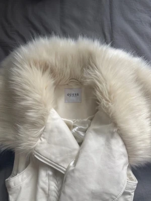 Vintage Vit pälsväst från Guess - Säljer en snygg vit pälsväst från Guess med stor fluffig fuskpälskrage. Västen har dragkedja framtill, två fickor med dragkedja och ett elegant mönster i tyget. Perfekt för att lyfta din outfit med en lyxig touch.