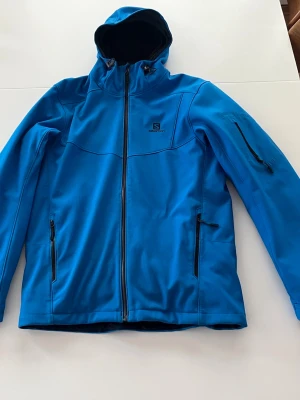 Blå skidjacka från Salomon - Blå strl M, är i gott skick men har en lite svart fläck på armbågen. Inget som syns i helhet. Jackan har liftkorts ficka, snölås samt en tillhörande fleece som går att fästa och ta loss med en knapp i nacken 