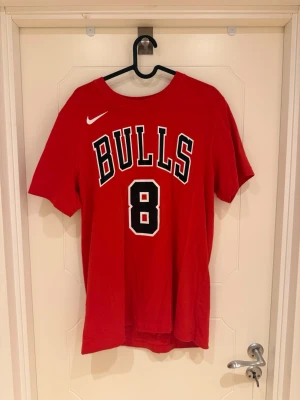 Zach Lavine Bulls T-Shirt - Säljer en röd t-shirt från Nike med Chicago Bulls-tryck och siffran 8 på bröstet. T-shirten har korta ärmar, rund halsringning och är tillverkad i mjuk bomull. Perfekt för dig som gillar basket och vill ha en sportig look.                                                                        Modellen är 185cm och väger 82kg.