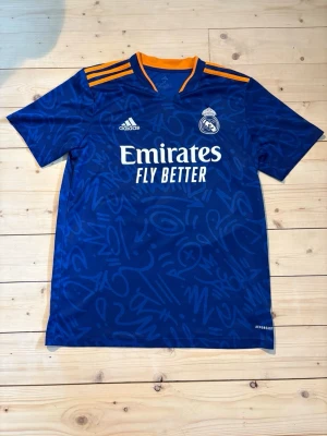 Blå Real Madrid matchtröja Adidas - Säljer en snygg bortatröja från Real Madrid säsongen 2021/2022 med Vinícius Júnior på ryggen.  ✅ Bra skick – inga fläckar på själva tröjan ℹ️ Mindre märke på siffran bak, bör gå bort i tvätten 👕 Bekväm och perfekt för match eller vardag  Hör av dig vid frågor!