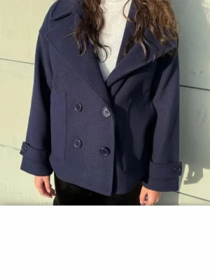 Mörkblå skepparkavaj med bred krage - Stilren mörkblå skepparkavaj med dubbelknäppning och bred krage. Jackan har stora knappar framtill och dekorativa knappar vid ärmsluten. Klassisk rak passform och tillverkad i ett tjockare tyg som håller värmen. Perfekt för dig som gillar tidlös och trendig stil.