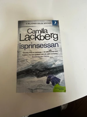 Isprinsessan av Camilla Läckberg - En spännande pocketbok, 'Isprinsessan' av Camilla Läckberg. Omslaget är grått och vitt med ismotiv och blå text. Boken är tryckt på papper och tillhör serien om polisen Patrik Hedström och författaren Erica Falck i Fjällbacka.