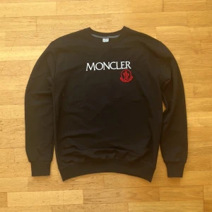 Svart Moncler sweatshirt - Svart sweatshirt från Moncler med vit logotyptext och röd broderad patch på bröstet. Klassisk rund halsringning, ribbade muddar vid ärmslut och nederkant. Perfekt för en clean och stilren look.