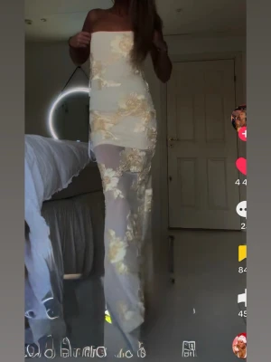 Bella barnett Klänning  - Bella barnett, Säljer en elegant långklänning i vit mesh med guldiga blommiga broderier. Klänningen är axelbandslös och har en figurnära passform som ger en snygg siluett. Perfekt för dig som vill sticka ut med en glamorös look.