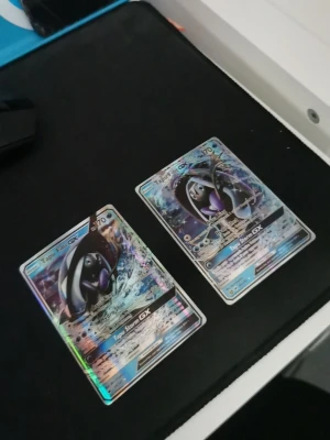 Två Tapu Fini GX Pokémonkort - Säljer två Tapu Fini GX samlarkort från Pokémon. Båda korten har holografisk yta med färger i blått, lila och silver. Perfekt för dig som vill utöka din samling med snygga och unika GX-kort. Kortens baksida har klassiskt Pokémon-motiv.
