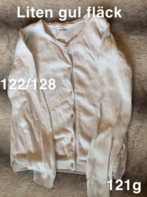 Vit kofta med guldknappar - Vit kofta med rund halsringning och snygga guldknappar framtill. Långärmad modell med ribbade muddar vid ärmslut och nederkant. Perfekt att slänga över en t-shirt för en enkel och stilren look.🌸 Liten gul fläck. Storlek 122/128