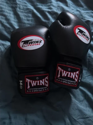 Svarta boxningshandskar Twins Special - Säljer ett par svarta boxningshandskar från Twins Special, storlek 14 oz. Handskarna har kardborreknäppning och tydliga loggor på ovansidan och vid handleden. Tillverkade i slitstarkt konstläder, perfekta för boxning eller thaiboxning.