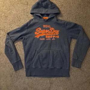 Blå Superdry hoodie med tryck - Snygg blå hoodie från Superdry med orange tryck på bröstet och japanska tecken. Klassisk känguruficka framtill och huva. Perfekt för en avslappnad streetwear-look.
