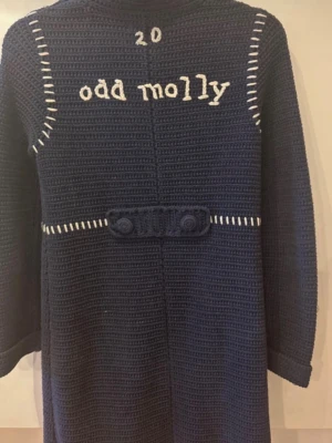 Mörkblå stickad kappa Odd Molly - Mörkblå stickad kappa från Odd Molly med vita detaljer och broderad logga på ryggen. Kappan har knappar framtill, två fickor och dekorativa sömmar. Långärmad och med en snygg krage. Perfekt för dig som vill ha en unik och mysig stil.