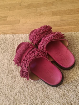 Rosa fransiga sandaler från Joseph Copenhagen  - Säljer ett par coola rosa?! sandaler från Joseph Copenhagen med breda remmar täckta av långa fransar. Sandalerna har en öppen tå och en platt sula i svart. Perfekta för dig som vill sticka ut med din stil och gillar unika detaljer.