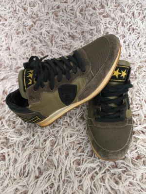 Gröna Phillipe model skor - Snygga sneakers i olivgrönt tyg och mocka med svarta detaljer och svarta skosnören. Tungan har en patch med tre gula stjärnor. Yttersulan är i gummi och skon har en låg profil. Perfekta för dig som gillar streetwear och unika detaljer. Har tyvärr inte kvar original kartongen! Bra skick, dom är välanvända men omhändertagna💕 dom är i mocka och har lite camouflage inslag med snygga ”militär” detaljer!!