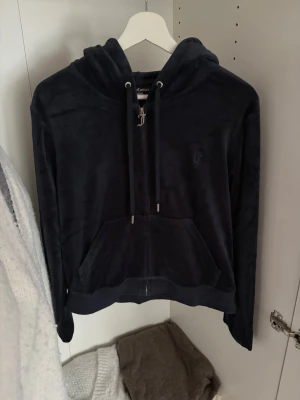 Mörkblå ziptröja med luva (velourhoodie) Juicy Couture - Säljer en mörkblå velourhoodie från Juicy Couture med dragkedja, huva med snören. Klassisk JC-logga broderad på bröstet och en snygg metall-detalj på dragkedjan. Perfekt för en chill och trendig look.