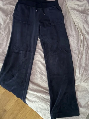 Juicy Couture, Mörkblå mjukisbyxor med raka ben - Säljer ett par mörkblå mjukisbyxor från Juicy Couture med raka ben och bred resår i midjan. Byxorna har dragsko och stora fickor framtill. Materialet är mjukt och skönt, perfekt för chill dagar. Diskret broderad logga på ena fickan.