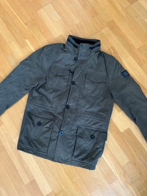 Olivgrön parkas med fickor - Stilren olivgrön parkas med fyra stora fickor framtill och hög krage. Jackan har knappstängning och en patch på ena ärmen. Materialet ser ut att vara slitstarkt och passar perfekt för kyligare dagar.
