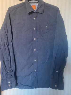 Blå linneskjorta från BOSS - Snygg blå skjorta från BOSS i linne med klassisk krage och bröstficka. Skjortan har vita knappar framtill och på manschetterna samt lång ärm. Perfekt för en stilren och avslappnad look.