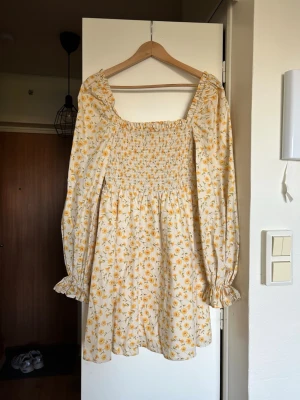 Blommig kortklänning med långa ärmar - Supersöt kortklänning med smock upptill och långa puffärmar. Klänningen är oanvänd med gult blommönster, har volangdetaljer vid ärmslut och en insydd underklänning. 