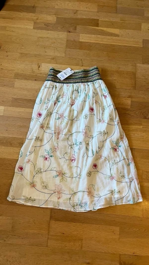 Långkjol - Säljer en vit långkjol med blommigt mönster i pastellfärger och bred smock i midjan. Kjolen har en luftig och flowy passform, perfekt för varma dagar. Materialet känns lätt och bekvämt mot huden.