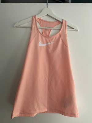 Nike Dri-Fit ljusrosa träningslinne - Säljer ett ljusrosa träningslinne från Nike med Dri-Fit material som andas. Linnet har brottarrygg och en vit Nike-logga på bröstet. Perfekt för gymmet eller löpning när du vill ha något svalt och bekvämt.