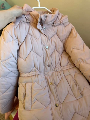 Ljusrosa dunjacka med huva - Säljer en ljusrosa dunjacka med huva och snyggt mönstrad quiltning. Jackan har dragkedja och tryckknappar framtill, två fickor och resår i midjan för en schysst passform. Perfekt för kalla vinterdagar och riktigt mysig att bära.