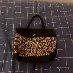 Shoppingväska med leopardmönster - Snygg shoppingväska med svart botten och handtag samt en stor panel i leopardmönster på framsidan. Väskan har en rymlig form och är perfekt för att bära med sig allt man behöver. Materialet känns slitstarkt och väskan har en liten metallbricka som detalj framtill.