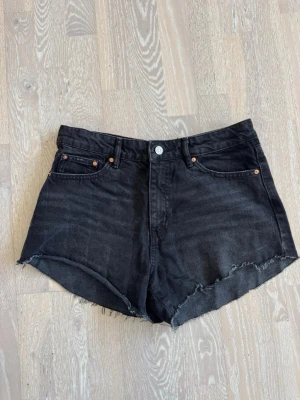 Svarta jeansshorts med rå kant - Snygga svarta jeansshorts med hög midja och rå, fransig kant. Klassisk femficksmodell med knapp och dragkedja framtill. Perfekta till sommaren för en avslappnad och trendig look! Ej använda☺️