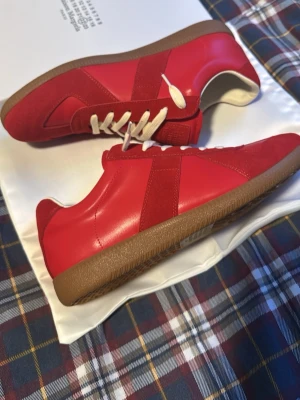Röda sneakers Maison Margiela - Snygga röda sneakers från Maison Margiela med ovandel i läder och mocka, vita skosnören och klassisk gummisula i brunt. Modellen har en retroinspirerad design med lågt skaft och diskreta detaljer. Perfekta för dig som vill sticka ut med stil.