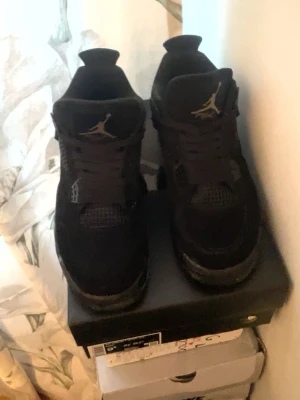 Svarta Air Jordan 4 Retro sneakers - Säljer ett par helsvarta Air Jordan 4 Retro sneakers med klassisk siluett och Jumpman-logga på plösen. Skorna har meshdetaljer, plastvingar och är tillverkade i mocka och textil. Perfekta för dig som gillar streetwear och vill ha en ikonisk look.
