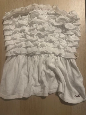 Vit bandeau topp från Hollister - Säljer en supersöt vit bandeau topp från Hollister med volangdetaljer och rynkad överdel. Toppen är axelbandslös och har en flowy passform nedtill. Perfekt för varma dagar och enkel att styla med jeans eller kjol. Ta kontakt om du har några frågor 