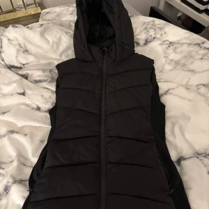 Svart lång dunväst med huva - Snygg svart dunväst med huva och dragkedja framtill från zara. Den är endast använd 1-3 gånger och får ingen användning av den längre. Finns ett litet hål (se bild 2) men det går lätt att sy ihop och inget man märker av. 