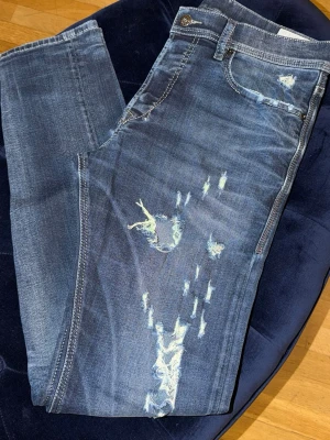 Blå slitna jeans med distressed detaljer - Säljer ett par blå jeans med snygga slitningar och distressed detaljer på framsidan. Jeansen har klassisk femficksmodell och raka ben. Perfekta för en avslappnad och trendig look. Materialet är mjukt denim som ger en skön känsla.