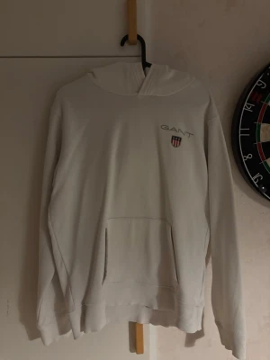 Vit hoodie från GANT med logga - Snygg vit hoodie från GANT med broderad logga och sköld på bröstet. Klassisk känguruficka framtill och ribbade muddar vid ärmslut och nederkant. Tillverkad i mjukt material för maximal komfort. Perfekt för en avslappnad och stilren look.