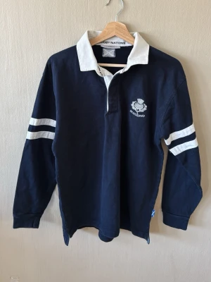 Mörkblå rugbytröja Rugby Nations - Klassisk mörkblå rugbytröja från Rugby Nations med vit krage och vita ränder på ärmarna. Broderad Skottland-logga på bröstet och lång ärm. Tillverkad i kraftig bomull, perfekt för sport eller chill.