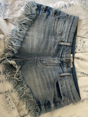 Blå jeansshorts med fransar - Säljer ett par blå jeansshorts med råa fransade kanter och klassisk femficksdesign. Shortsen har normal passform och är tillverkade i bomull. Perfekta för sommaren och ger en avslappnad vibe. Skulle säga att storleken är mellan 25/26. Säljer pågrund av för stora❤️