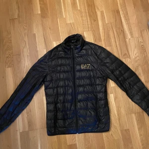 Svart dunjacka från Emporio Armani - Snygg svart dunjacka till vår/sommar från EA7 Emporio Armani med guldfärgad logga på bröstet. Köpte den på rea förra vintern men har inte använt så mycket. Nypris 1300kr