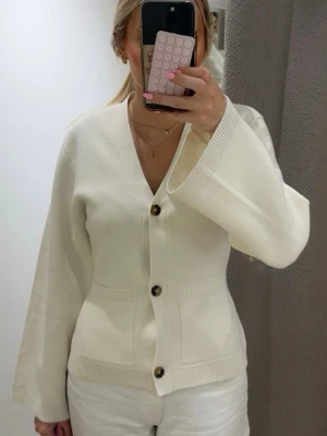 Krämvit kofta med vida ärmar - Stilren krämvit kofta med v-ringning och stora bruna knappar framtill. Koftan har vida ärmar, ribbade muddar och två fickor framtill. Perfekt för en avslappnad men snygg look.