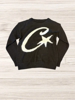 Svart stickad Corteiz tröja - Cool svart stickad tröja med stor vit C och stjärna på framsidan. Tröjan har rund halsringning och långa ärmar. Perfekt för dig som gillar grafiska tryck och vill sticka ut lite extra. Materialet känns mjukt och bekvämt. Skriv till mig vid några funderingar.