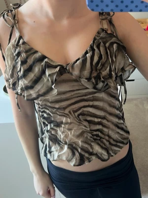 Zebramönstrad linnetopp med knyt - Säljer en zebramönstrad linnetopp med tunna axelband och knyt på axlarna. Toppen har volangdetaljer framtill och är i ett lätt, luftigt material. Perfekt för dig som vill sticka ut med ett djurmönster och gillar trendiga detaljer.