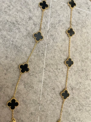 Van cleef halsband  - Snyggt halsband med guldfärgad kedja och flera svarta klöverformade hängen. Klöverhänget har en tunn guldram som ger en exklusiv känsla. Perfekt accessoar för dig som gillar stilrena och trendiga smycken.