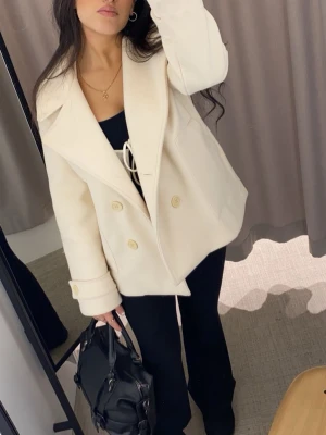 Krämvit skepparkavaj med stora slag - Stilren krämvit skepparkavaj med dubbelknäppning och stora slag. Jackan har breda manschetter med knappdetalj och är tillverkad i ett mjukt, tjockt tyg som ger en lyxig känsla. Perfekt för dig som vill ha en klassisk och trendig look.